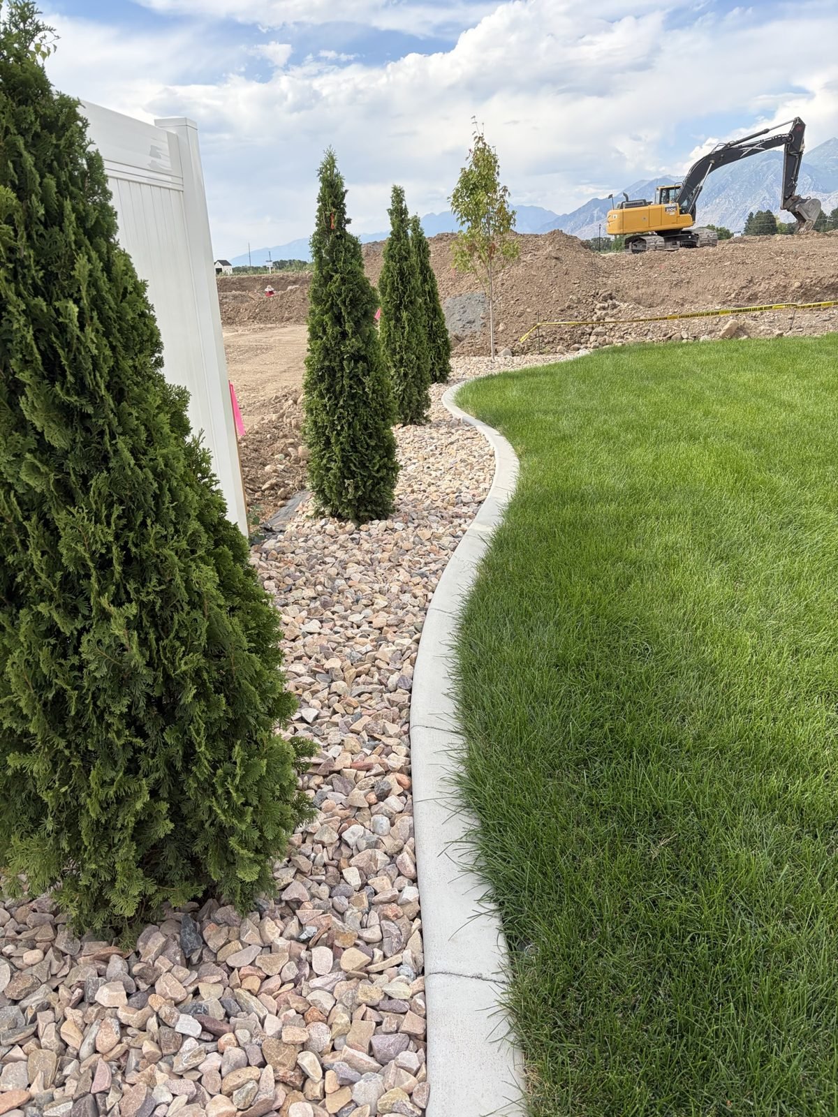 Evergreens, river rock and concrete curbing next to new sod on a Roots Landscaping install in Utah County